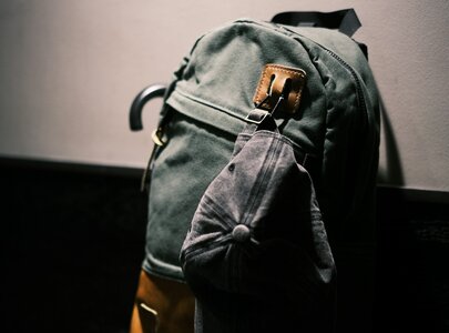 Ein grüner Rucksack hängt an einer Wand, mit einer braunen Unterseite. Eine graue Baseballkappe ist an einem der Riemen des Rucksacks befestigt. Die Umgebung ist dunkel, wodurch die Konturen des Rucksacks deutlich hervorstechen.