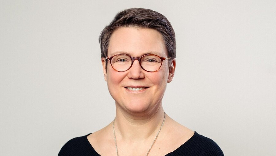Eine lächelnde Person mit kurzen Haaren und einer modischen Brille trägt ein schwarzes Oberteil. Sie steht vor einem hellen Hintergrund, ihre Pose vermittelt Freundlichkeit und Offenheit.