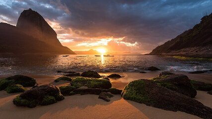 Ein Sonnenuntergang über dem Wasser, mit orangefarbenen und goldenen Tönen. Im Vordergrund liegen durch das Wasser glitschige, moosbedeckte Steine, während eine große Bergformation auf der linken Seite sichtbar ist. Der Himmel ist teilweise bewölkt und strahlt warmes Licht aus.