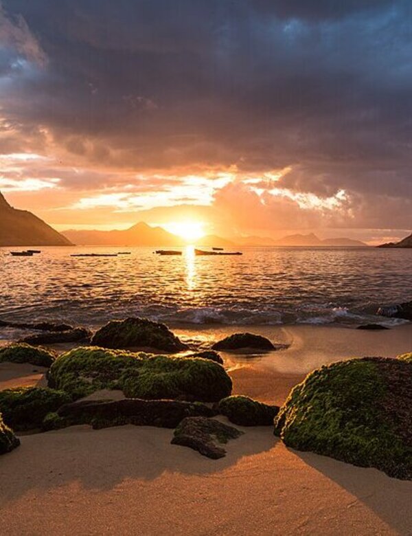 Ein Sonnenuntergang über dem Wasser, mit orangefarbenen und goldenen Tönen. Im Vordergrund liegen durch das Wasser glitschige, moosbedeckte Steine, während eine große Bergformation auf der linken Seite sichtbar ist. Der Himmel ist teilweise bewölkt und strahlt warmes Licht aus.