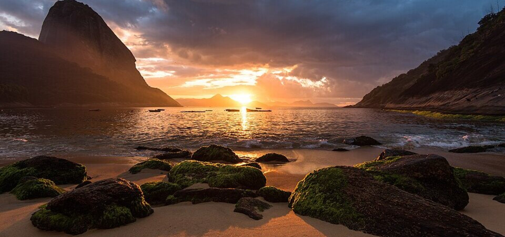 Ein Sonnenuntergang über dem Wasser, mit orangefarbenen und goldenen Tönen. Im Vordergrund liegen durch das Wasser glitschige, moosbedeckte Steine, während eine große Bergformation auf der linken Seite sichtbar ist. Der Himmel ist teilweise bewölkt und strahlt warmes Licht aus.