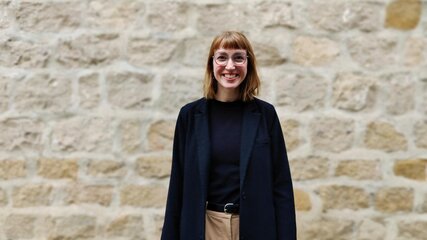 Eine lächelnde Frau mit kurzen, braunen Haaren und Brille steht vor einer steinernen Wand. Sie trägt einen schwarzen Blazer und ein schwarzes Oberteil sowie helle, bequeme Hosen. Ihr freundlicher Ausdruck vermittelt Offenheit und Selbstbewusstsein.