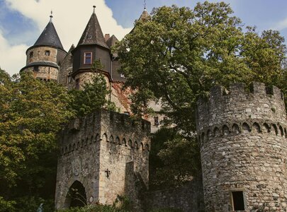 Eine alte Burgruine, umgeben von Bäumen, mit mehreren Türmen und Zinnen. Der Hauptturm hat eine spitze Dachform, während das Eingangstor von einer massiven Steinmauer flankiert wird. Der Himmel ist teils bewölkt, und die gesamte Szenerie vermittelt einen historischen Eindruck.