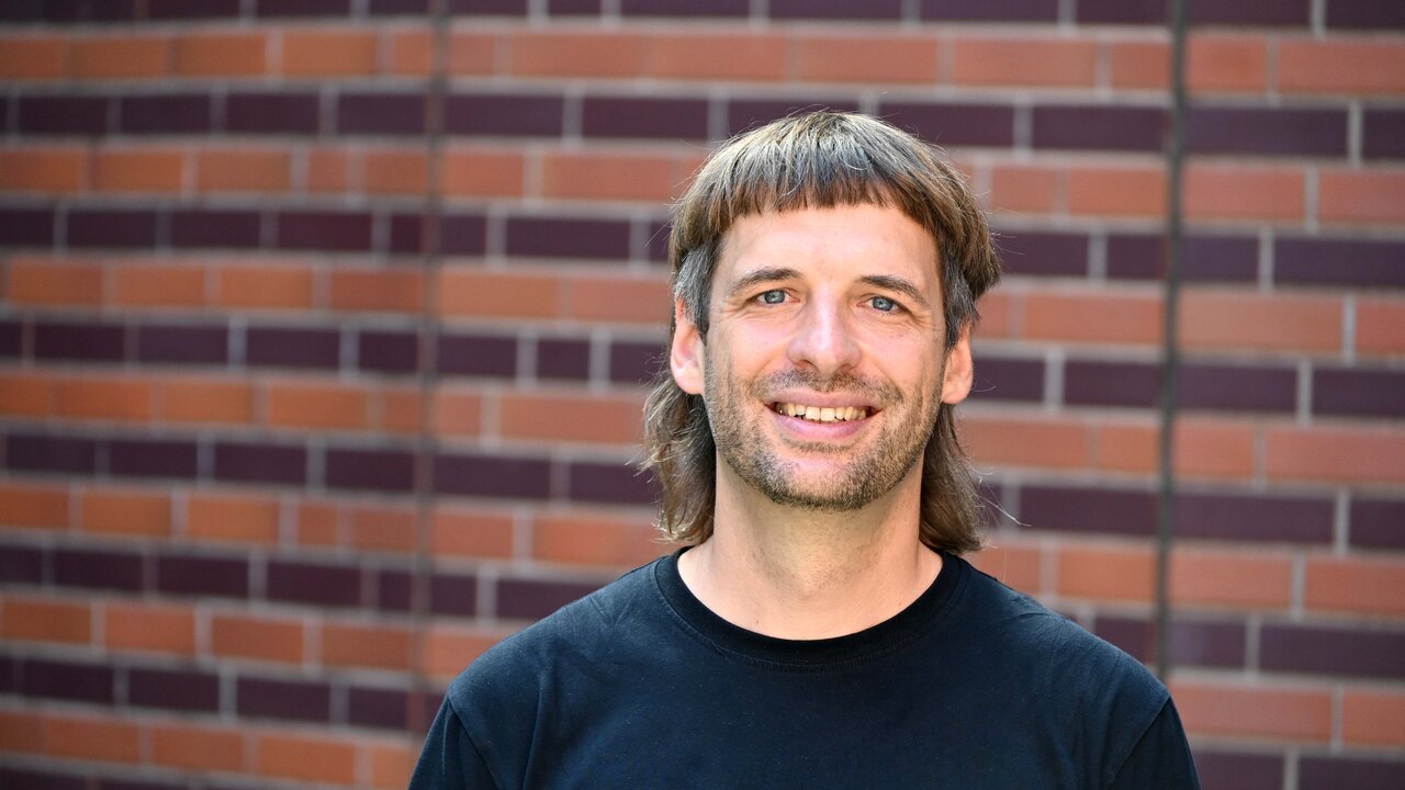 Ein Mann mit einem schwarzen T-Shirt lächelt vor einer braun-rot gemusterten Mauer. Er hat mittellange, strähnige Haare, die zu einem Schnitt mit Pony gestylt sind. Der Hintergrund hebt die freundliche Ausstrahlung des Mannes hervor.
