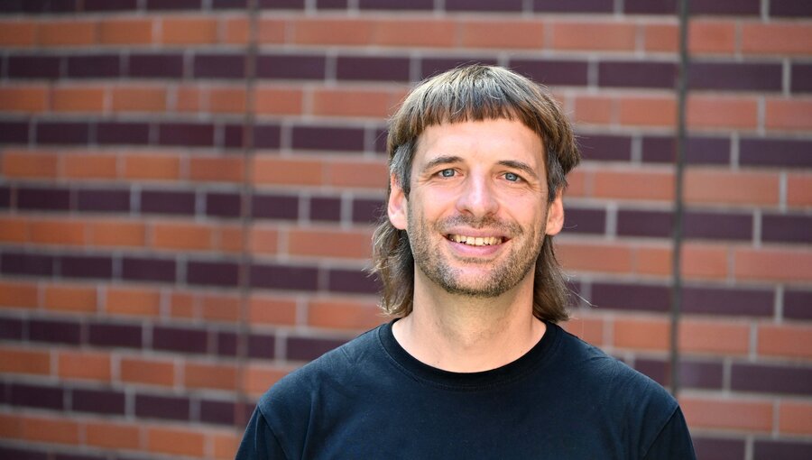 Ein Mann mit einem schwarzen T-Shirt lächelt vor einer braun-rot gemusterten Mauer. Er hat mittellange, strähnige Haare, die zu einem Schnitt mit Pony gestylt sind. Der Hintergrund hebt die freundliche Ausstrahlung des Mannes hervor.