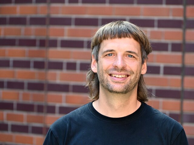 Ein Mann mit einem schwarzen T-Shirt lächelt vor einer braun-rot gemusterten Mauer. Er hat mittellange, strähnige Haare, die zu einem Schnitt mit Pony gestylt sind. Der Hintergrund hebt die freundliche Ausstrahlung des Mannes hervor.