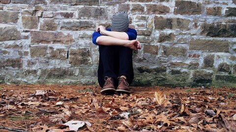 Eine sitzende Person in einem blauen Oberteil und einer gestreiften Mütze hockt mit angewinkelten Beinen an einer Steinmauer. Der Boden ist mit trockenen Blättern und Nadelstreu bedeckt, was eine melancholische Atmosphäre vermittelt.