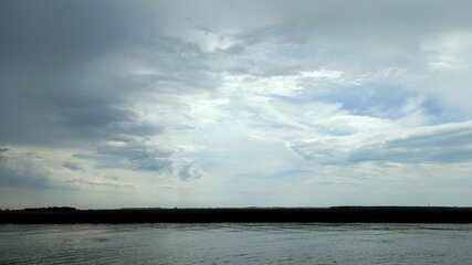 Eine ruhige Wasserfläche spiegelt einen bewölkten Himmel wider. Über dem Wasser sind graue und weiße Wolken mit blauen Stellen zu sehen, während am Horizont eine grüne Landschaft angedeutet wird. Das Bild vermittelt eine friedliche, aber melancholische Stimmung.