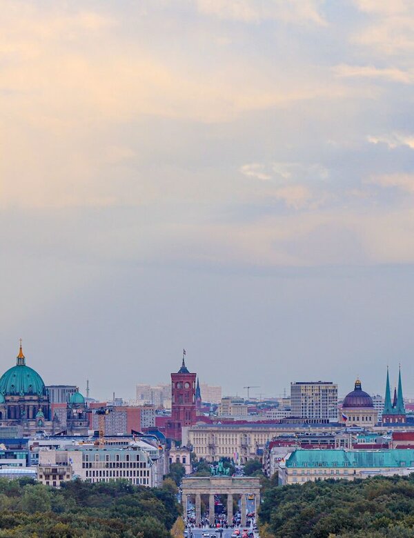 Ein Blick auf die Berliner Skyline mit dem Fernsehturm, der das Stadtbild dominiert. Im Hintergrund sind die Kuppel der Berliner Kirche und verschiedene historische Gebäude sichtbar. Vor der Stadt erstreckt sich eine Allee, die von Bäumen gesäumt ist.
