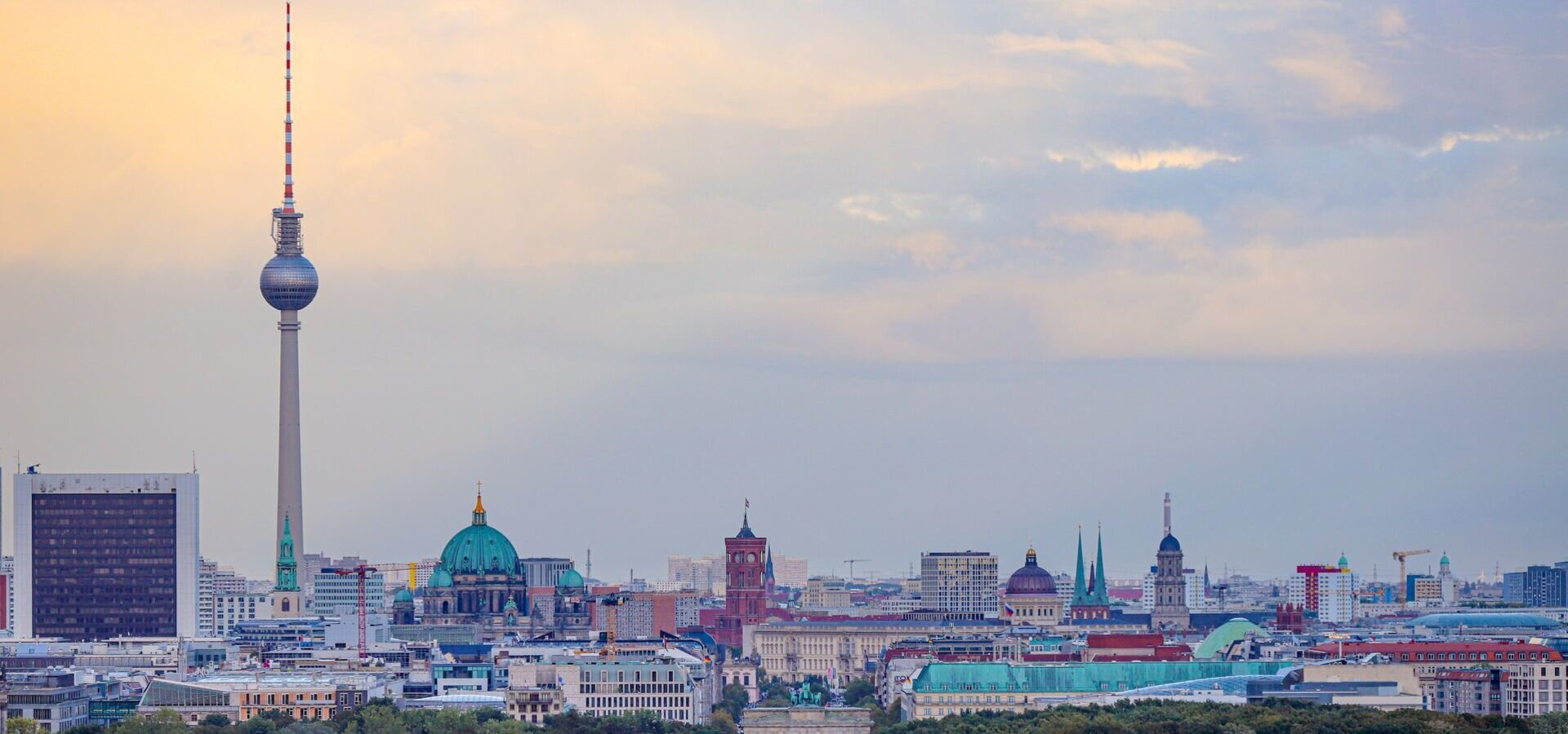 Ein Blick auf die Berliner Skyline mit dem Fernsehturm, der das Stadtbild dominiert. Im Hintergrund sind die Kuppel der Berliner Kirche und verschiedene historische Gebäude sichtbar. Vor der Stadt erstreckt sich eine Allee, die von Bäumen gesäumt ist.