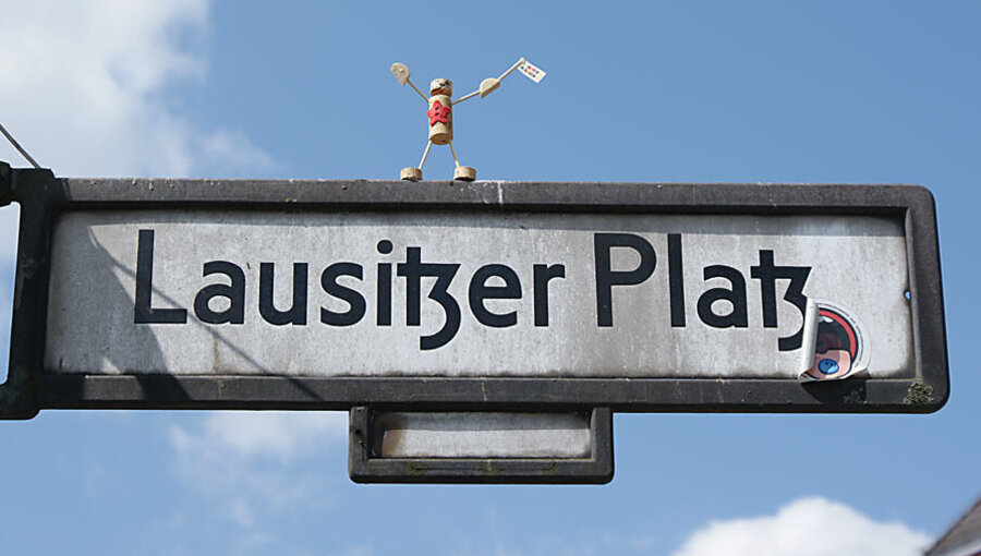 Ein Straßenschild zeigt den Namen „Lausitzer Platz“. Oben auf dem Schild steht eine kleine Figur, die ein Schild in der Hand hält und fröhlich posiert. Der Himmel ist blau mit einigen Wolken.