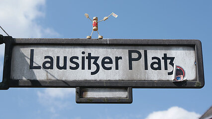 Ein Straßenschild zeigt den Namen „Lausitzer Platz“. Oben auf dem Schild steht eine kleine Figur, die ein Schild in der Hand hält und fröhlich posiert. Der Himmel ist blau mit einigen Wolken.
