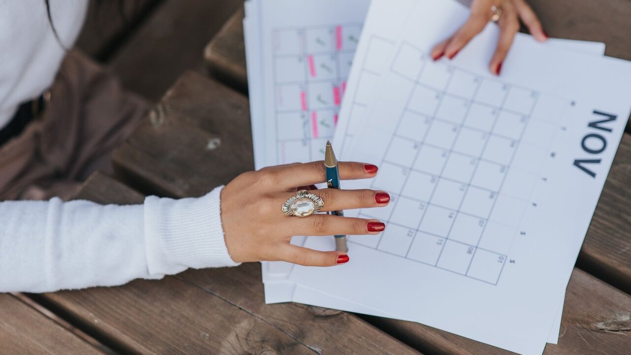 Eine Hand mit einem auffälligen Ring hält einen Stift und zeigt auf einen Kalender, der den Monat November anzeigt. Im Hintergrund liegen weitere Blätter. Die Umgebung besteht aus einer Holzoberfläche.