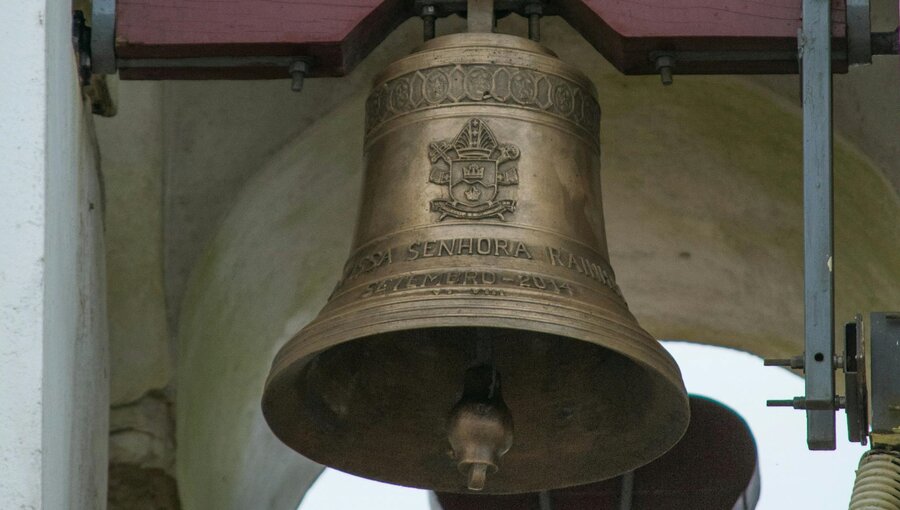 Eine große, goldene Glocke hängt in einem Glockenturm. Auf der Glocke sind Wappen und Schriftzüge zu sehen, die vermutlich religiöse Bedeutung haben. Der Hintergrund ist unscharf und zeigt die Struktur des Turms.