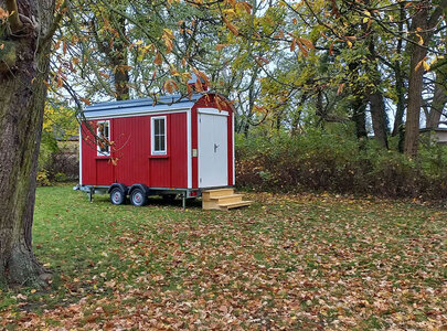 Ein rotes, kleines Mobilheim steht auf einer Wiese, umgeben von Laubbäumen. Vor dem Haus befindet sich eine Treppe und ein Schild mit der Aufschrift "Offene Kirche". Der Boden ist mit buntem Herbstlaub bedeckt.