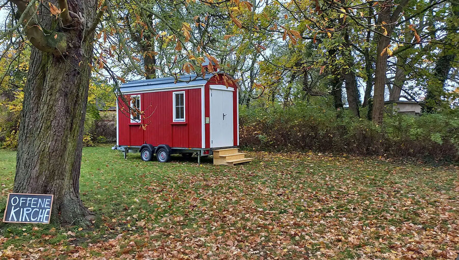 Ein rotes, kleines Mobilheim steht auf einer Wiese, umgeben von Laubbäumen. Vor dem Haus befindet sich eine Treppe und ein Schild mit der Aufschrift "Offene Kirche". Der Boden ist mit buntem Herbstlaub bedeckt.