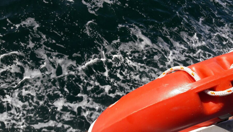 Ein orangefarbener Rettungsring liegt auf dem Rand eines Bootes, während dunkles Wasser mit kleinen Wellen und Schaum spritzt. Das Bild vermittelt ein Gefühl von Sicherheit auf dem Wasser.