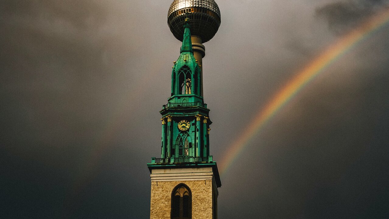 Ein hoher Turm mit einer markanten Kugelspitze steht vor einem dramatischen, bewölkten Himmel. Regenbogen färben den Himmel hinter dem Turm, während die Uhr an der Vorderseite des Turms die Zeit anzeigt.