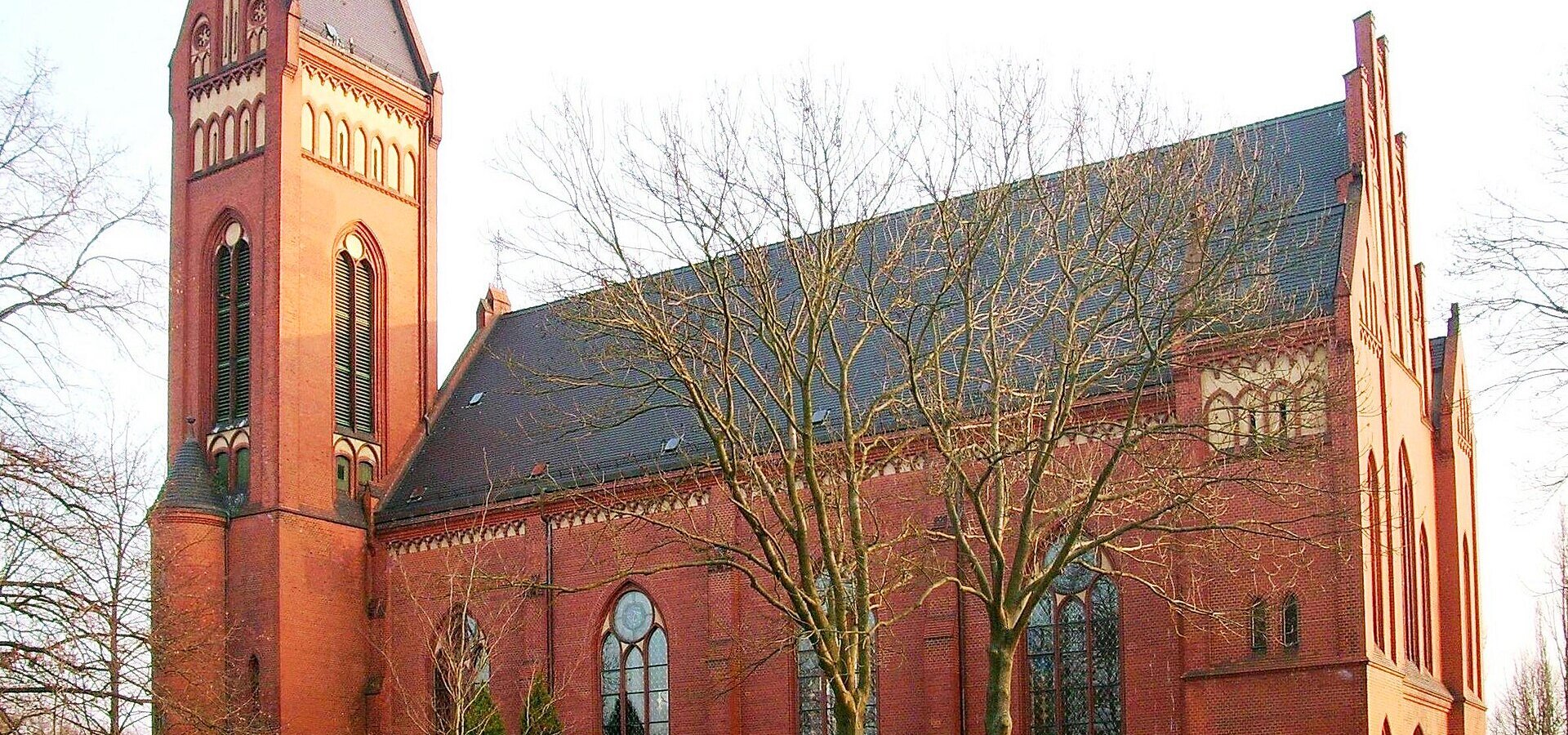 Ein roter Backsteinkirche mit hohen Fenstern und einem markanten Turm, umgeben von kahlen Bäumen. Die Architektur ist gotisch inspiriert und die Fassade zeigt filigrane Details. Die Umgebung ist weitläufig und ruhig, was der Kirche eine friedliche Ausstrahlung verleiht.