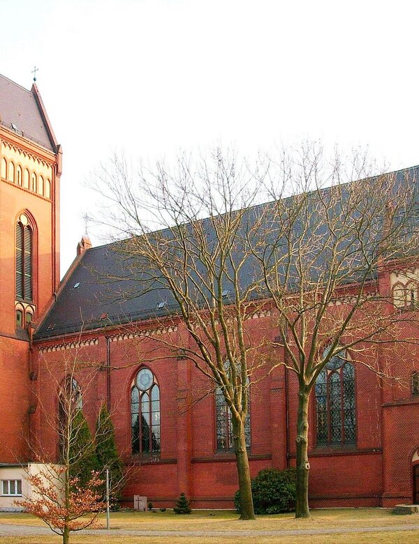 Ein roter Backsteinkirche mit hohen Fenstern und einem markanten Turm, umgeben von kahlen Bäumen. Die Architektur ist gotisch inspiriert und die Fassade zeigt filigrane Details. Die Umgebung ist weitläufig und ruhig, was der Kirche eine friedliche Ausstrahlung verleiht.