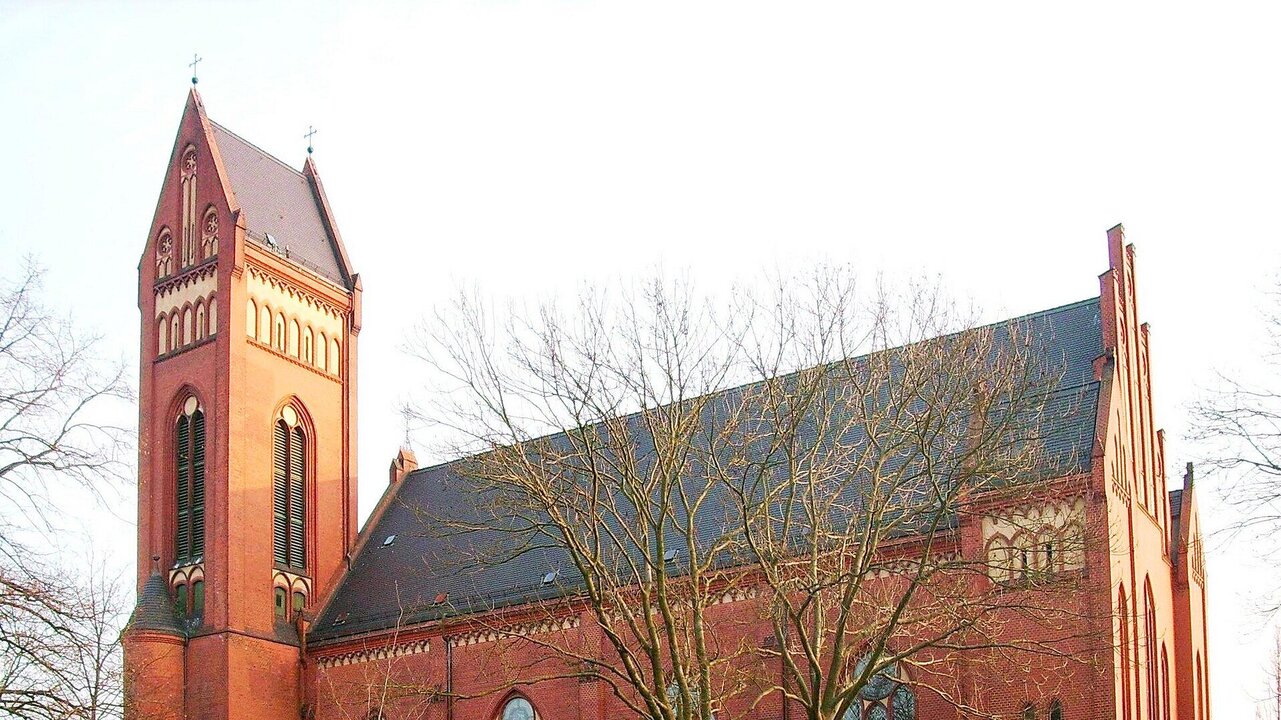 Ein roter Backsteinkirche mit hohen Fenstern und einem markanten Turm, umgeben von kahlen Bäumen. Die Architektur ist gotisch inspiriert und die Fassade zeigt filigrane Details. Die Umgebung ist weitläufig und ruhig, was der Kirche eine friedliche Ausstrahlung verleiht.
