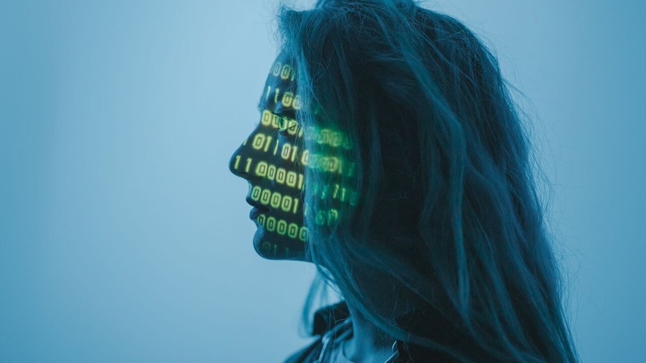 Ein Profil einer Person mit langen Haaren, deren Gesicht mit grünen binären Zahlen (Nullen und Einsen) projiziert ist. Der Hintergrund ist in sanften Blautönen gehalten. Die Darstellung symbolisiert eine Verbindung zwischen Mensch und Technologie.