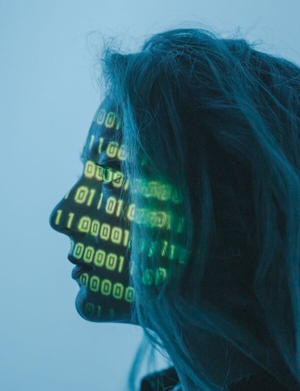 Ein Profil einer Person mit langen Haaren, deren Gesicht mit grünen binären Zahlen (Nullen und Einsen) projiziert ist. Der Hintergrund ist in sanften Blautönen gehalten. Die Darstellung symbolisiert eine Verbindung zwischen Mensch und Technologie.