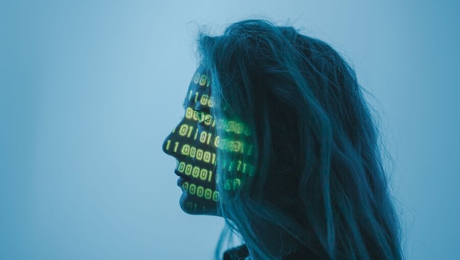 Ein Profil einer Person mit langen Haaren, deren Gesicht mit grünen binären Zahlen (Nullen und Einsen) projiziert ist. Der Hintergrund ist in sanften Blautönen gehalten. Die Darstellung symbolisiert eine Verbindung zwischen Mensch und Technologie.