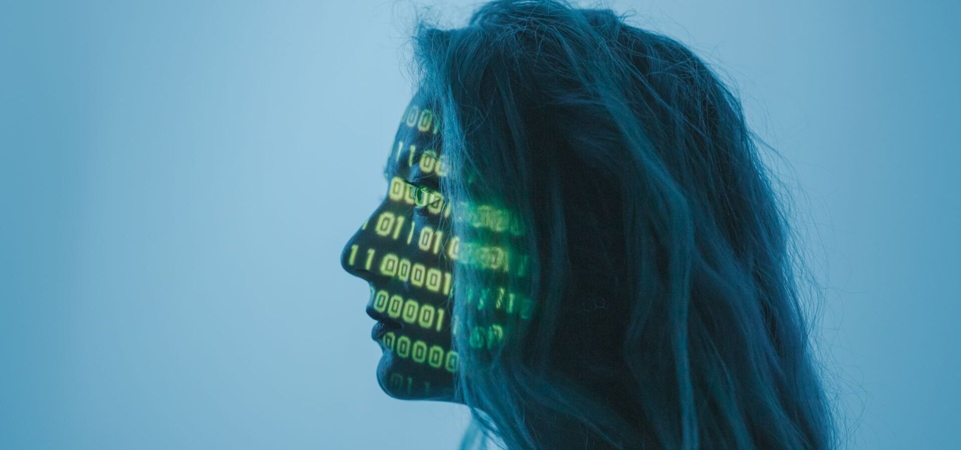 Ein Profil einer Person mit langen Haaren, deren Gesicht mit grünen binären Zahlen (Nullen und Einsen) projiziert ist. Der Hintergrund ist in sanften Blautönen gehalten. Die Darstellung symbolisiert eine Verbindung zwischen Mensch und Technologie.