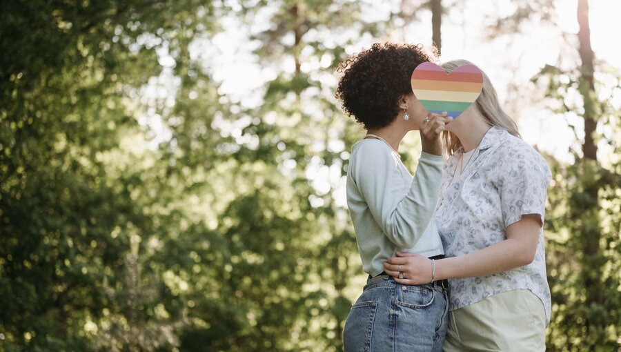 Zwei Frauen stehen sich gegenüber in einer natürlichen Umgebung. Sie halten sich an der Taille und küssen sich, während sie miteinander in einem liebevollen Moment verbunden sind. Im Hintergrund sind Bäume und Sonnenlicht sichtbar, das eine warme Atmosphäre schafft.