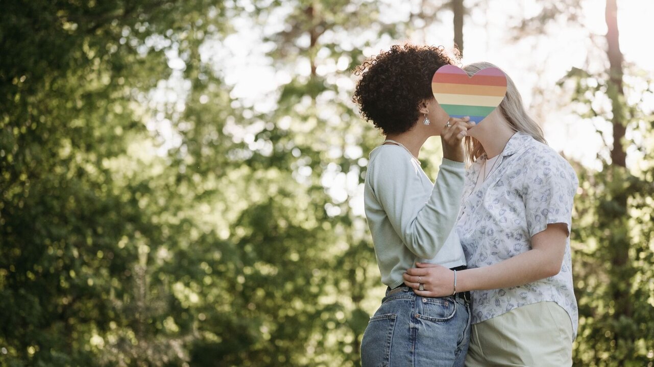 Zwei Frauen stehen sich gegenüber in einer natürlichen Umgebung. Sie halten sich an der Taille und küssen sich, während sie miteinander in einem liebevollen Moment verbunden sind. Im Hintergrund sind Bäume und Sonnenlicht sichtbar, das eine warme Atmosphäre schafft.
