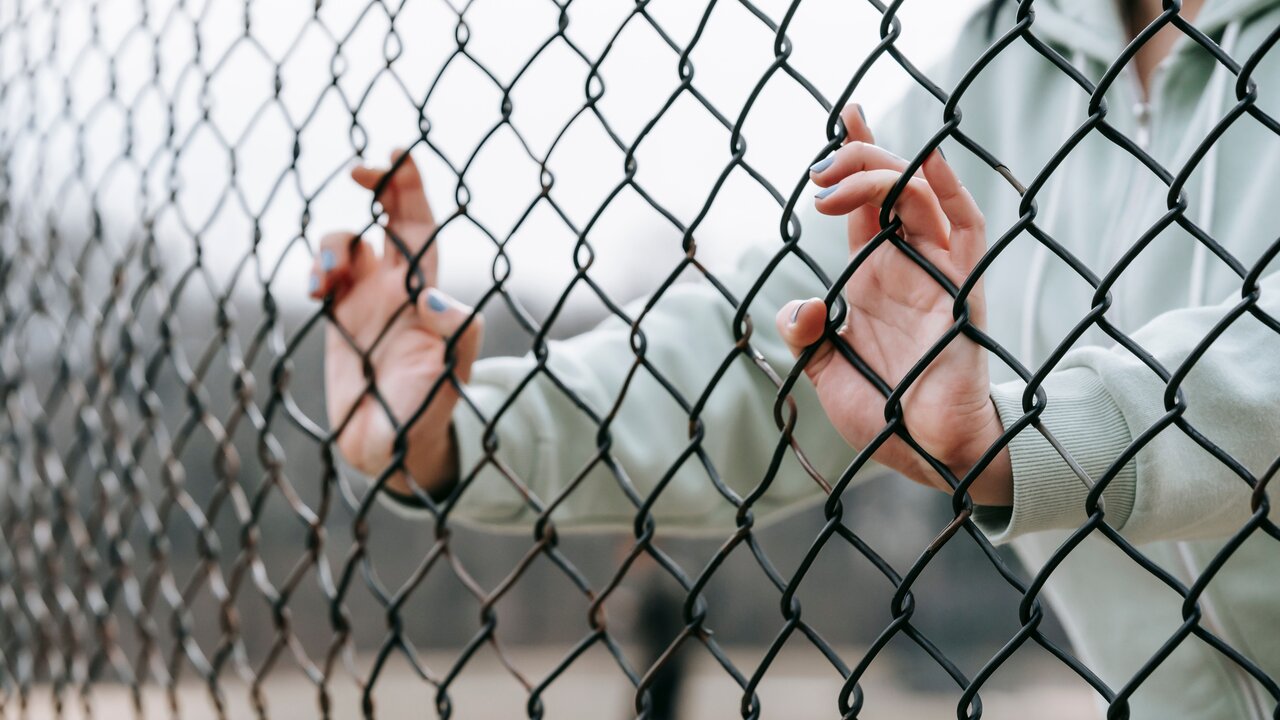 Eine Nahaufnahme von Händen, die an einem Maschendrahtzaun greifen. Die Person trägt einen grauen Pullover und der Fokus liegt auf den fingern, die sich durch das Netz hindurch drängen. Im Hintergrund sind unscharfe Strukturen zu sehen.