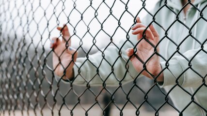 Eine Nahaufnahme von Händen, die an einem Maschendrahtzaun greifen. Die Person trägt einen grauen Pullover und der Fokus liegt auf den fingern, die sich durch das Netz hindurch drängen. Im Hintergrund sind unscharfe Strukturen zu sehen.