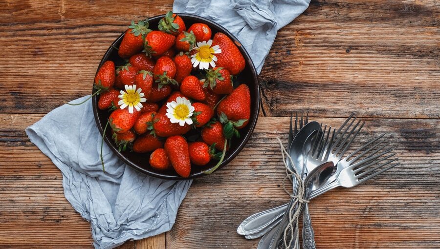 Eine Schüssel mit frischen, roten Erdbeeren, garniert mit weißen Margeriten, steht auf einem rustikalen Holztisch. Daneben liegen mehrere Besteckteile, einschließlich Gabeln und Löffel, die mit twine zusammengebunden sind.
