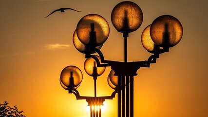 Der Sonnenuntergang beleuchtet mehrere Laternen mit runden Glaskugeln, die sanft leuchten. Im Hintergrund fliegt ein Vogel silhouettenhaft durch den orangefarbenen Himmel.