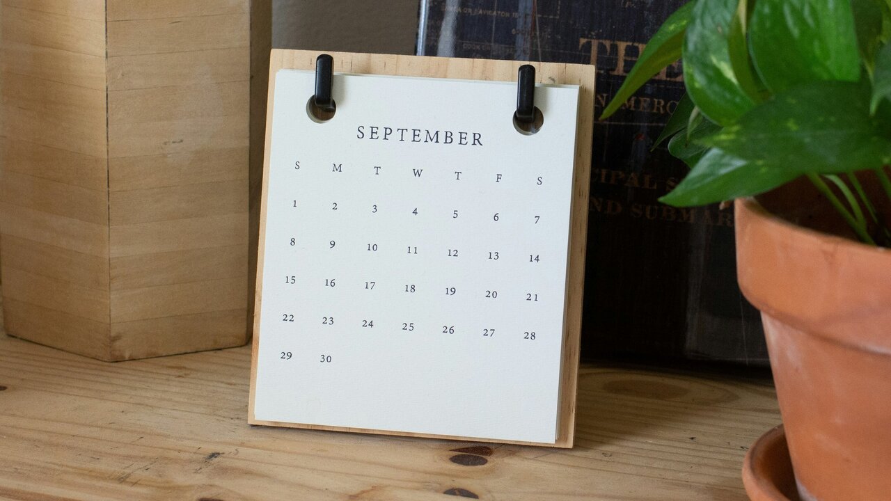 Ein Tischkalender für den Monat September steht auf einem Holztisch. Er zeigt die Tage von Montag bis Sonntag sowie die Zahlen für den Monat. Neben dem Kalender steht eine Topfpflanze.
