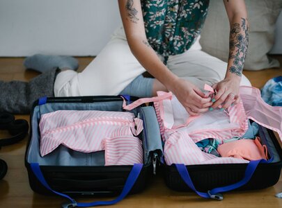 Eine Person sitzt auf dem Boden und packt einen Koffer. Der Koffer ist offen und enthält verschiedene Kleidungsstücke, darunter gestreifte Blusen und Badeanzüge. Die Person hat sichtbare Tattoos und trägt eine bunte Bluse. Um sie herum liegen weitere Kleidungsstücke.