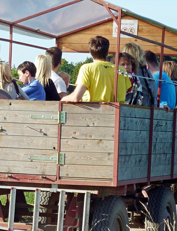 Eine Gruppe von Menschen sitzt in einem offenen Anhänger, der von einem Traktor gezogen wird. Die Szenerie spielt auf einem landwirtschaftlichen Gelände mit grüner Umgebung. Einige Personen lächeln und unterhalten sich, während der Anhänger auf einem schmalen Weg fährt.