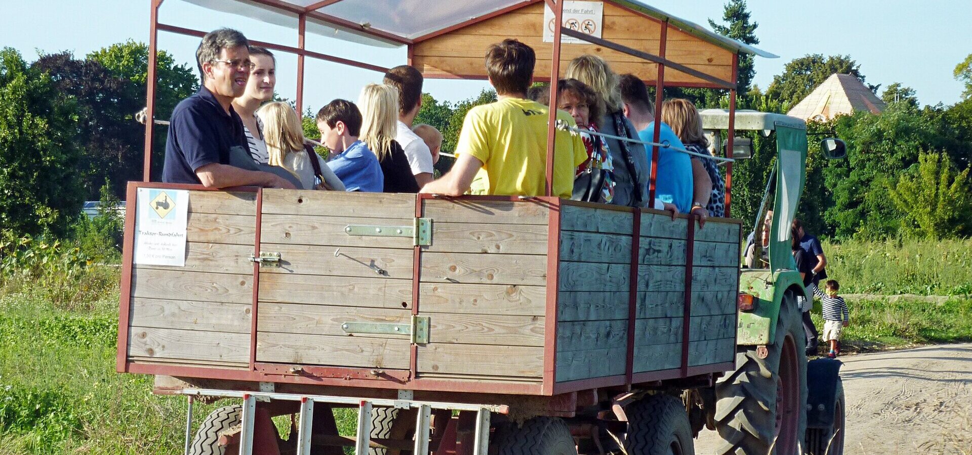 Eine Gruppe von Menschen sitzt in einem offenen Anhänger, der von einem Traktor gezogen wird. Die Szenerie spielt auf einem landwirtschaftlichen Gelände mit grüner Umgebung. Einige Personen lächeln und unterhalten sich, während der Anhänger auf einem schmalen Weg fährt.