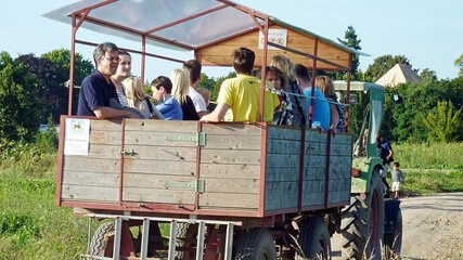 Eine Gruppe von Menschen sitzt in einem offenen Anhänger, der von einem Traktor gezogen wird. Die Szenerie spielt auf einem landwirtschaftlichen Gelände mit grüner Umgebung. Einige Personen lächeln und unterhalten sich, während der Anhänger auf einem schmalen Weg fährt.