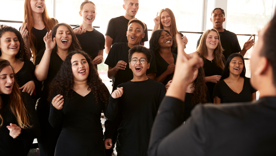 Eine Gruppe von Sängern steht in einem Probenraum, trägt schwarze Kleidung und singt gemeinsam. Einige Sänger lächeln und zeigen Freude an der Musik. Im Vordergrund ist ein Aufsicht führender Dirigent zu sehen, der die Gruppe anleitet.