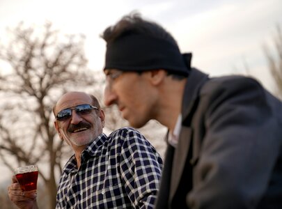 Zwei Männer sitzen im Freien und unterhalten sich. Der Mann links trägt eine Sonnenbrille und hält ein Glas mit rotem Getränk. Der Mann rechts hat ein Stirnband an und schaut zur Seite. Im Hintergrund sind einige Bäume sichtbar.