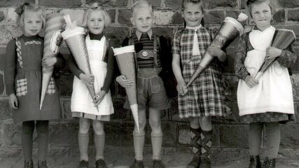 Fünf Kinder stehen vor einer Steinmauer, alle sind festlich gekleidet und tragen große, bunte Schultüten. Zwei Mädchen in Hüten und Kleider, ein Junge in einer Latzhose sowie ein weiteres Mädchen in schickem Outfit zeigen Vorfreude. Die Szene strahlt Freude auf den Schulanfang aus.
