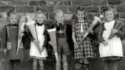 Fünf Kinder stehen vor einer Steinmauer, alle sind festlich gekleidet und tragen große, bunte Schultüten. Zwei Mädchen in Hüten und Kleider, ein Junge in einer Latzhose sowie ein weiteres Mädchen in schickem Outfit zeigen Vorfreude. Die Szene strahlt Freude auf den Schulanfang aus.