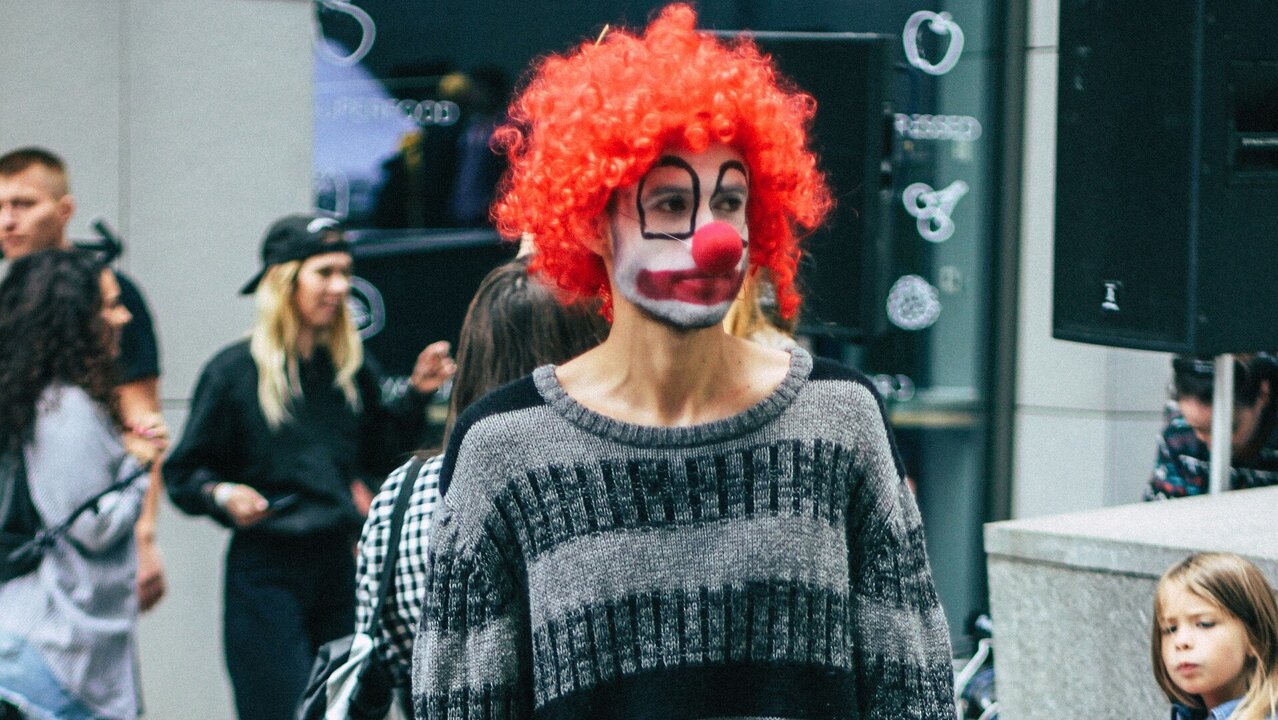 Eine Person im Clownskostüm mit roter Perücke hält ein handbeschriebenes Plakat, auf dem steht: „Life is a Fucking Circus“. Die Person trägt einen gestreiften Pullover und steht in einer urbanen Umgebung.