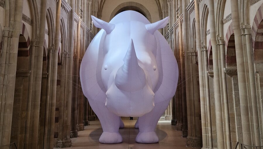 Ein riesiges, aufblasbares Nashorn steht in einem großen, gewölbten Raum mit hohen Säulen. Es ist weiß und hat eine charakteristische Form mit einem gebogenen Horn. Die Umgebung vermittelt eine beeindruckende, historische Atmosphäre.