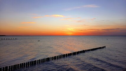 Ein malerischer Sonnenuntergang über dem Meer, mit warmen Farben von Orange und Lila, die den Himmel färben. Sanfte Wellen spiegeln das Licht wider, während ein Holzsteg im Wasser verläuft. Die ruhige Stimmung vermittelt ein Gefühl von Frieden und Harmonie.