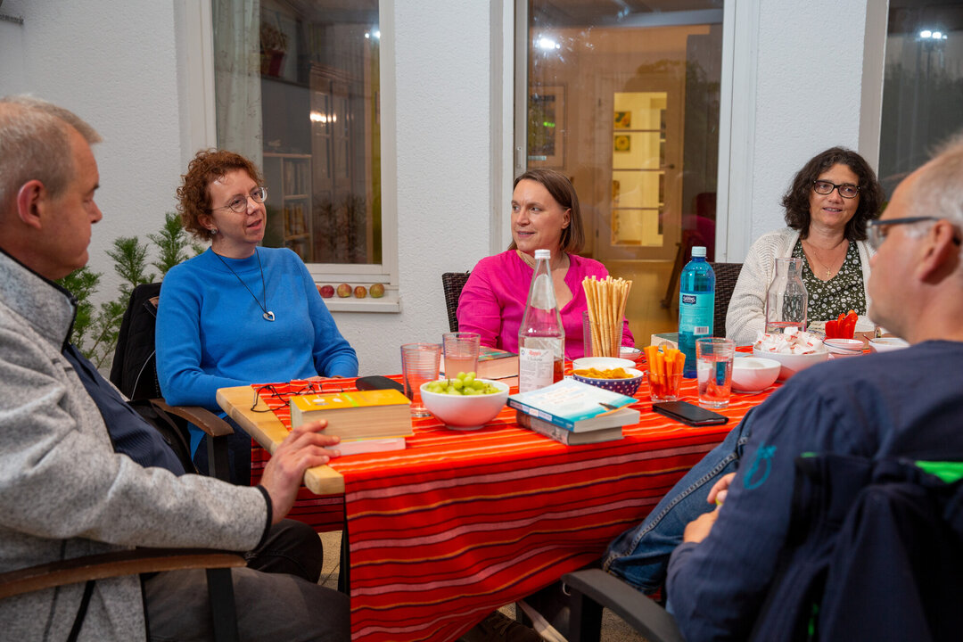 Sechs Personen sitzen an einem Tisch, der mit einem roten Tischdecken und verschiedenen Getränken sowie Snacks gedeckt ist. Sie scheinen sich anregend zu unterhalten und genießen die gemeinsame Zeit. Die Atmosphäre wirkt freundlich und entspannt.