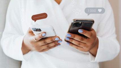 Eine Person in einem weißen Bademantel hält zwei Smartphones in den Händen. Auf dem Bildschirm eines Handys sind eine Sprechblase und eine Anzeige für 1000 Herzen zu sehen, was auf Kommunikation und Interaktion hinweist.