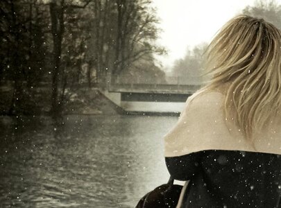 Eine Frau mit langen, blonden Haaren steht am Ufer eines Gewässers. Der Schnee fällt leicht, während sie nachdenklich in die Ferne blickt. Im Hintergrund sind Bäume und eine Brücke sichtbar, die eine ruhige, winterliche Atmosphäre schaffen.